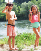 Ein Junge in Main Design Lean Boardshorts für Jungen hält einen Stock und steht barfuß neben einem lächelnden Mädchen in einem rosafarbenen und türkisfarbenen Badeanzug an einem Fluss mit üppigem Grün und Bäumen hinter ihnen.