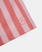 Nahaufnahme der rosa und rot vertikal gestreiften Lean Boardshorts für Jungen von Main Design mit aufgenähtem kreisförmigem Logo mit stilisiertem "A" in der Nähe des Saums. Das schnell trocknende Material bietet UPF 50+ Schutz auf hellem Grund.