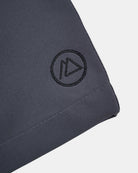 Eine Nahaufnahme der dunkelgrauen Lean Boardshorts für Jungen von Main Design zeigt ein aufgesticktes, rundes Logo mit einem stilisierten "A" in einem Dreieck in der Nähe des Saums; das Material ist schnell trocknend und bietet UPF 50+ Sonnenschutz.