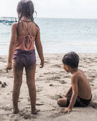 Zwei kleine Kinder spielen an einem Sandstrand; das Mädchen, das die Lena Mädchen Badeanzug von Main Design mit UPF 50+ trägt, steht mit Blick auf das Meer, während der Junge in der Nähe unter einem bedeckten Himmel im Sand sitzt, während die Wellen ruhig anrollen.