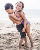 Zwei kleine Kinder, ein Mädchen und ein Junge, lächeln, während sie an einem Sandstrand spielen und sich umarmen. Das Mädchen trägt den Lena Mädchen Badeanzug von Main Design, mit dem Meer und dem Himmel im Hintergrund. Beide sind mit Sand bedeckt und sehen glücklich aus.