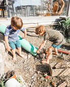 Zwei kleine Jungen, barfuß in Main Design Lean Boardshorts für Jungen mit blauen Mustern, spielen draußen auf einem unbefestigten Hang mit Steinen und einer grünen Poolnudel als Rampe, während Spielzeug und Gartengeräte verstreut herumliegen.
