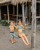 Zwei Kinder spielen auf einer Holzschaukel am Strand. Das Mädchen sitzt in einem grünen Badeanzug, während der Junge in der Nähe in der Lean Boardshorts für Jungen von Main Design steht. Im Hintergrund sind eine strohgedeckte Hütte und Holzmöbel zu sehen.