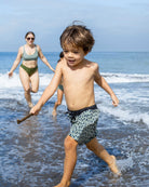 Ein kleiner Junge in Main Design Lean Boardshorts für Jungen läuft an einem sonnigen Strand, hält einen Stock und lächelt, während seine Boardshorts mit UPF 50+ schützen. Hinter ihm spielen eine Frau und ein anderes Kind in den ruhigen Wellen des Ozeans.