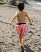 Ein Junge mit kurzen dunklen Haaren, der Lean Boardshorts für Jungen von Main Design trägt, läuft barfuß über einen Sandstrand in Richtung Wasser, den Rücken zur Kamera.
