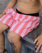 Ein Kind in rosa-weiß gestreiften Lean Boardshorts für Jungen von Main Design mit UPF 50+ und 4-Way Stretch sitzt auf Holz, Sand an den nackten Beinen. Die Badeshorts haben ein kleines rundes Logo auf einem Bein.