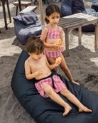 Zwei Jungen in passenden rosa-weißen Lean Boardshorts für Jungen von Main Design mit 4-Wege-Stretch genießen den Strand - einer sitzt mit sandigen Beinen auf einem schwarzen Sitzsack, während der andere in der Nähe steht und Obst hält. Hinter ihnen stehen Holztische und Stühle.