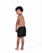 Ein junges Kind mit kurzen dunklen Haaren, barfuß und mit Main Design Lean Boardshorts für Jungen und einer Halskette bekleidet, blickt vor einem schlichten weißen Hintergrund über seine Schulter.