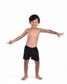 Ein Junge trägt die Lean Boardshorts für Jungen von Main Design und eine bunte Perlenkette, steht barfuß vor weißem Hintergrund, lächelt und streckt die Arme aus - ideal für Abenteuer dank schnell trocknender Boardshorts.
