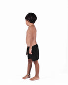 Ein Junge mit kurzen dunklen Haaren steht barfuß, trägt Main Design Lean Boardshorts für Jungen, eine Halskette und ein Fußkettchen und blickt nach links auf einen schlichten weißen Hintergrund.