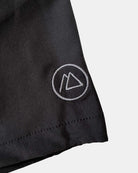 Eine Nahaufnahme der Nolan Boardshorts für Jungen von Main Design in Schwarz hebt ein kleines weißes, rundes Logo mit zwei sich überlappenden Dreiecken in der Nähe des Saums hervor und verfügt über 4-Way-Stretch für zusätzlichen Komfort.