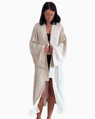 Eine Person steht barfuß vor einem schlichten weißen Hintergrund und trägt einen langen, beigen, strukturierten Kimono Amazona von Main Design über einem schwarzen Outfit. Das Gewand hat weite Ärmel und ist vorne offen, wobei die Person die Ränder sanft festhält. Der Kopf ist leicht geneigt.