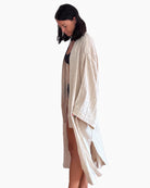 Eine Person mit schulterlangem dunklem Haar steht barfuß und trägt ein langes, beiges, fließendes Gewand über einem schwarzen Outfit, das an den Kimono Amazona von Main Design erinnert. Der Kopf der Person ist vor einem schlichten weißen Hintergrund leicht nach unten geneigt.