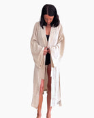 Eine Person mit langen, dunklen Haaren schaut nach unten, während sie den Gürtel eines beigen, langärmeligen Kimono Amazona-Gewandes von Main Design bindet. Sie ist barfuß und trägt unter dem Gewand vor einem schlichten weißen Hintergrund ein schwarzes Outfit aus weichem Material.