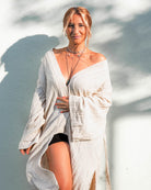 Eine Person steht lächelnd vor einer sonnenbeschienenen Wand in Main Designs Kimono Amazona. Das leichte, fließende Gewand fällt elegant in seiner Einheitsgröße und umarmt die Figur, während die Person mit mehreren im Licht schimmernden Halsketten vortritt, die einen Schatten auf die strukturierte Oberfläche werfen.