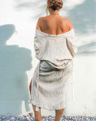 Eine Frau mit einem Haarknoten steht barfuß im Freien vor einer strukturierten weißen Wand. Sie trägt den „Kimono Amazona“ von Main Design, ein schulterfreies Überwurf aus Leinen-Baumwoll-Mischung. Ihr Schatten wird auf die Wand geworfen und sie steht auf Gras mit kleinen Steinen.