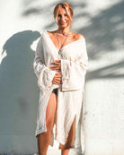 Eine Person, die barfuß auf einem Kiesweg vor einer hellen Wand steht, ist in den Kimono Amazona von Main Design gehüllt, ein lockeres, schulterfreies beiges Kleidungsstück aus Leinen-Baumwoll-Mischung. Sie ist mit mehreren Halsketten geschmückt, während Sonnenlicht und Schatten die entspannte Atmosphäre verstärken.