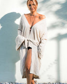 Eine Frau steht vor einer hellen Wand und trägt den Kimono Amazona von Main Design. Das lockere, schulterfreie Kleid ist aus weicher Leinen-Baumwoll-Mischung gefertigt. Sie trägt mehrere Halsketten und hat ihr Haar zu einem Dutt frisiert. Barfuß auf dem Kieselboden lächelt sie sanft.