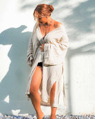 Barfuß auf einem Kiesweg vor einer hellen Wand präsentiert die Person den eleganten Kimono Amazona von Main Design. Teilweise schimmert schwarze Kleidung durch, die Haare sind zu einem Knoten gebunden, das sanfte Licht wirft sanfte Schatten.