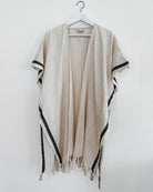 Der Kimono Naya von Main Design - ein beigefarbener, leicht gestreifter Kimono mit schwarzen Akzenten und Fransen - hängt auf einem weißen Bügel an einer schlichten weißen Wand. Ideal als stilvoller Strandüberwurf oder Sommerkimono.