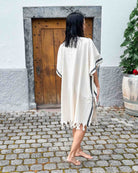 Eine Person mit nassen Haaren steht barfuß auf einem Kopfsteinpflasterweg vor einer Holztür. Sie trägt den Kimono Naya von Main Design - ein weißes, knielanges Kleidungsstück mit Fransen und schwarzen Streifen - während rechts eine große Topfpflanze steht.
