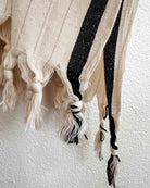 Eine Nahaufnahme des Kimono Naya von Main Design zeigt cremefarbene OEKO-TEX-zertifizierte Baumwolle mit schwarzen Streifen und geknoteten Fransen vor einer strukturierten weißen Wand.