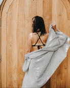 Eine Person mit nassen Haaren steht vor einer großen Holztür. Sie trägt ein schwarzes Bikinioberteil und hat den Kimono Sakura von Main Design, handgefertigt in der Türkei, um die Schultern geschlungen. Die Szene scheint im Freien zu sein.