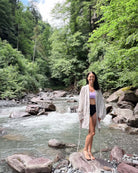 Eine Frau steht barfuß auf einem großen Felsen neben einem fließenden Fluss und lächelt friedlich in dem Kimono Sakura von Main Design, umgeben von üppig grünen Bäumen und genießt die Schönheit der Natur.