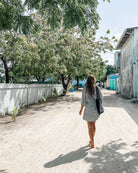 Eine Frau mit langen Haaren, barfuß und mit einer schwarzen Tasche in der Hand, schlendert in der Sonne einen von Bäumen und Gebäuden gesäumten Sandweg entlang. Sie trägt den Kimono Sakura von Main Design.