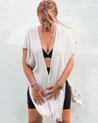 Eine hellbraunhaarige Frau trägt ein schwarzes Oberteil und den Kimono Saturn Cardigan mit offener Vorderseite von Main Design aus nachhaltigen Materialien in hellem Beige und dazu dunkle Shorts. Sie steht vor einer weißen Wand und trägt Halsketten, Armbänder und ein rosa Haargummi am Handgelenk.