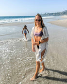 Eine Frau im Kimono Saturn von Main Design und mit Sonnenbrille geht am Strand entlang. Das leichte Design ihres Überwurfs fällt anmutig. Ein Kind im Badeanzug folgt ihr. Unter dem klaren Himmel plätschern Wellen an ihre Füße, umarmt von der nachhaltigen Schönheit der Natur.