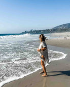 Eine Frau im Main Design Kimono Saturn aus nachhaltigen Materialien und Sandalen schlendert an einem Sandstrand entlang, während die Wellen anrollen. Der klare blaue Himmel gibt den Blick auf Hügel und ein paar Häuser in der Ferne frei.