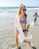 Eine Frau im Kimono Saturn und mit Sonnenbrille von Main Design schlendert am sonnigen Strand entlang, in dessen Nähe Wellen schwimmen, während hinter ihr ein Kind in einem weißen Oberteil und einer blauen Hose fröhlich im Wasser spielt.