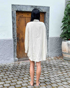 Eine barfuß gehende Person mit nassem, dunklem Haar steht auf einem Steinpflaster vor einer Holztür und trägt den cremefarbenen Kimono Solana von Main Design. Weiße und graue Wände und eine große Topfpflanze auf der rechten Seite vervollständigen die Szene.