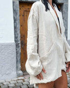 Eine Person mit nassen Haaren trägt den übergroßen Kimono Solana von Main Design in cremefarbener Farbe über schwarzer Kleidung. Sie steht im Freien auf Kopfsteinpflaster vor einer Holztür und einer weißen Wand, ihr Gesicht ist nicht zu erkennen.