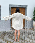 Eine Person steht barfuß und mit ausgestreckten Armen auf Kopfsteinpflaster und trägt den übergroßen Kimono Solana von Main Design. Sie steht vor einer in Stein gefassten Holztür, vor einer hellen Wand und neben einer Topfpflanze.