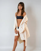 Eine Frau mit glattem braunem Haar und Pony steht vor einem schlichten, hellen Hintergrund, trägt einen schwarzen BH und Unterwäsche und hat den Kimono von Main Design Soleil - hergestellt aus 100% Baumwolle in nachhaltiger Produktion - über die Schultern gehängt.