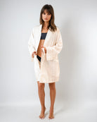 Eine Frau steht barfuß vor einem schlichten Hintergrund, trägt den Main Design Soleil Kimono - einen hellen, lockeren Bademantel aus 100 % Baumwolle - über einem schwarzen Bikini oder Unterwäsche und blickt mit neutralem Blick direkt in die Kamera.