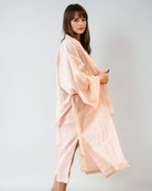 Eine Frau mit langen braunen Haaren steht seitlich und trägt den Main Design Sunset Stripes Kimono - ein lockeres, hellrosa und weiß gestreiftes Gewand mit weiten Ärmeln aus gekämmter Baumwolle - vor einem unifarbenen, hellen Hintergrund, ideal für einen entspannten Strandlook.