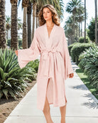 Eine Frau in dem rosa-weiß gestreiften Sunset Stripes Kimono von Main Design spaziert über einen sonnigen Bürgersteig mit Palmen und Grünpflanzen. Ihr ruhiges Selbstbewusstsein verkörpert mühelosen Strandstil.