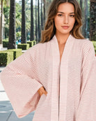 Eine Frau im Sunset Stripes Kimono von Main Design, mit weiten Ärmeln und einem hellen Rosaton, steht im Freien zwischen Palmen und gestutzten Büschen - ideal für einen entspannten Strandstil.