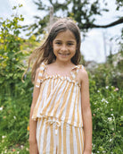 Ein junges Mädchen mit langen braunen Haaren lächelt draußen in einem grünen, blumenreichen Garten und trägt das Boho Hawaii Set von Main Design.