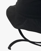 Eine Nahaufnahme eines schwarzen Kinder-Sonnenhuts mit breiter Krempe und zwei herabhängenden Kinnriemen. Der Hut aus umweltfreundlichem Stoff hat ein einfaches, schmuckloses Design und steht vor einem schlichten weißen Hintergrund.