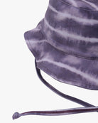 Eine Nahaufnahme eines Batik-Kinder-Sonnenhuts von Main Design in den Farben Schwarz, Dunkelgrau und Lila. Der Hut verfügt über einen verstellbaren Kinnriemen und eine breite Krempe. Der aus umweltfreundlichem Stoff gefertigte Stoff hat ein welliges, gestreiftes Muster für einen unverwechselbaren und lebendigen Look. Der Hintergrund ist schlicht weiß.