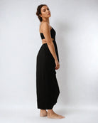 Eine Frau mit langem, welligem Haar steht barfuß vor einem schlichten weißen Hintergrund und trägt das Maxikleid Chasing the sun von Main Design. Sie blickt zur Seite, ihr Kopf ist leicht zur Kamera gedreht.