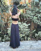 Eine Frau mit langen dunklen Haaren steht barfuß im Maxikleid Chasing The Sun Dress von Main Design auf einem Steinpfad, umgeben von üppigen grünen Pflanzen und Blättern.