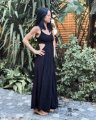 Eine Frau mit langen dunklen Haaren steht barfuß auf einem Steinpfad. Sie trägt das ärmellose Maxikleid Chasing The Sun Dress von Main Design mit seitlichen Ausschnitten und ist von üppigen grünen Pflanzen und Bäumen umgeben.