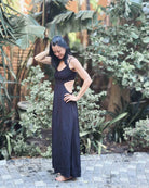 Eine Frau im Maxikleid „Chasing The Sun Dress“ von Main Design steht barfuß auf einem Kopfsteinpflasterweg inmitten üppigen Grüns, berührt sanft ihr Haar und lächelt sanft.