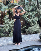 Eine Frau im langen schwarzen Maxikleid Chasing The Sun Dress von Main Design steht barfuß an einem kleinen Pool, die Hände im Haar, umgeben von üppigen grünen Pflanzen und Steinpflaster.