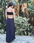 Eine Frau mit langem, dunklem Haar steht im Freien auf einem Steinweg und blickt abgewandt. Sie trägt das Main Design Maxikleid Chasing The Sun Dress mit gesmoktem Oberteil, umgeben von üppigen grünen tropischen Pflanzen.